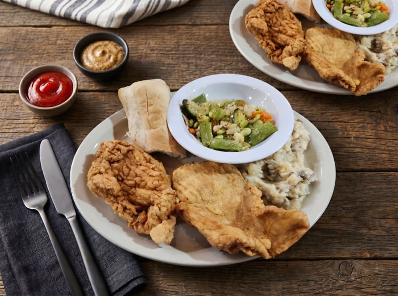 Two plates of fried chicken, mashed potatoes, green beans, and bread on a rustic wooden table.