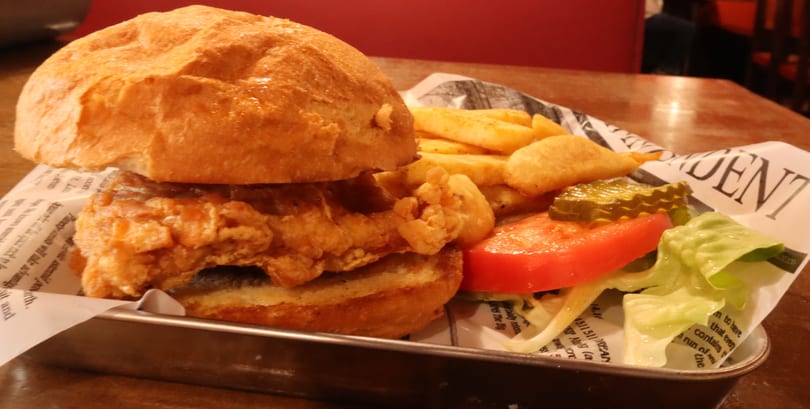 Crispy fried chicken sandwich with fries, tomato, lettuce, and pickles on a metal tray.