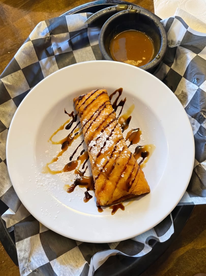 Golden-fried dessert chimichanga drizzled with chocolate and caramel, dusted with powdered sugar, served with dipping sauce.